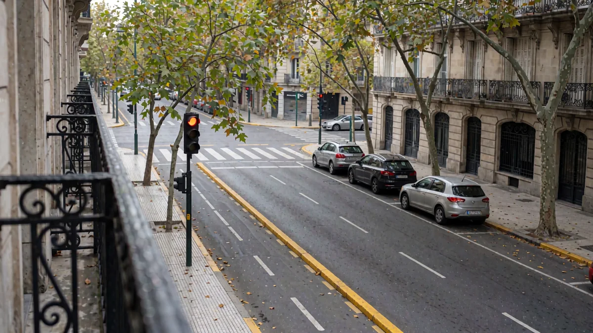 Cruce de una avenida porteña visto desde un balcón, con senda peatonal, ciclovía de separadores amarillos, dos autos estacionados y un semáforo en rojo entre tipas