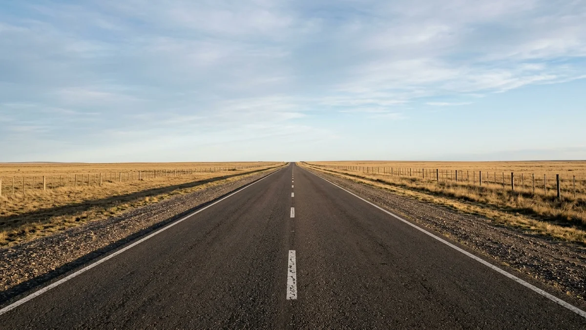 Ruta de dos carriles que cruza una llanura rural argentina al amanecer, con banquina de grava, alambrado paralelo y horizonte despejado.