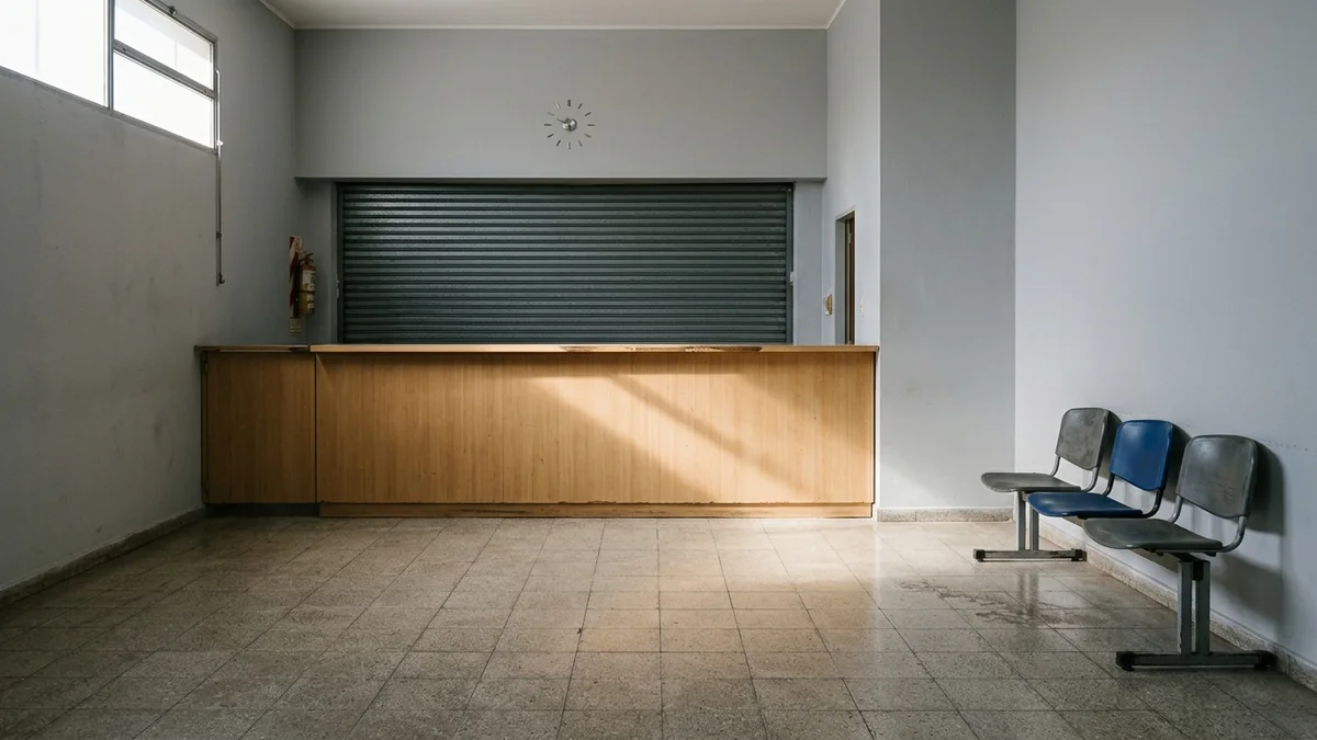 Hall vacío de una oficina pública con el mostrador de atención cerrado y la persiana metálica baja, iluminado por luz natural de la mañana.